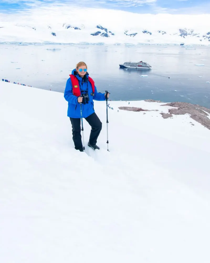 A traveller with walking poles on a snow slope in Antarctica, an expedition ship in the bay below