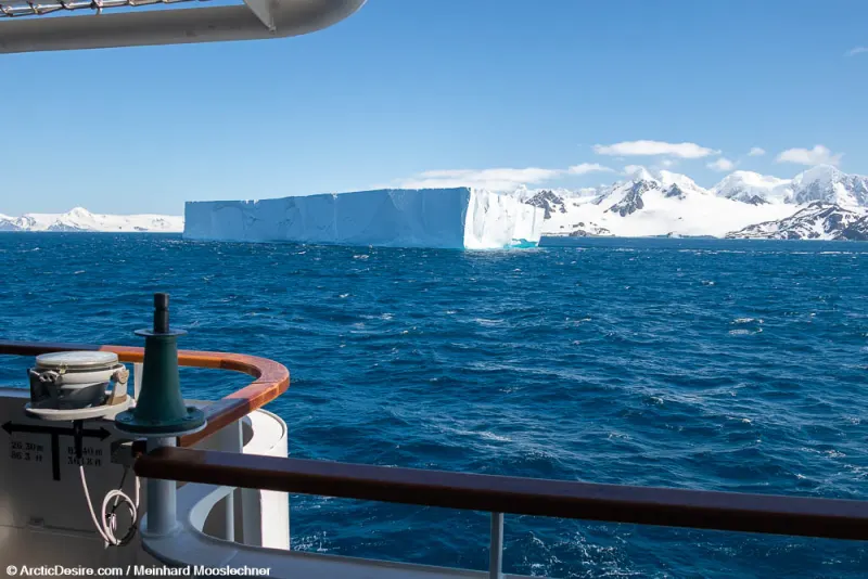 Blick von Deck eines Expeditionsschiffs auf einen Tafeleisberg vor verschneiten Bergen der Antarktis bei Sonnenschein