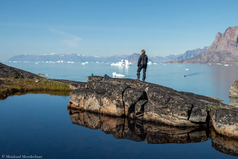 Ein Mann steht auf einem Felsen gegenüber von Uummannaq in Westgrönland, im Wasser treiben Eisberge