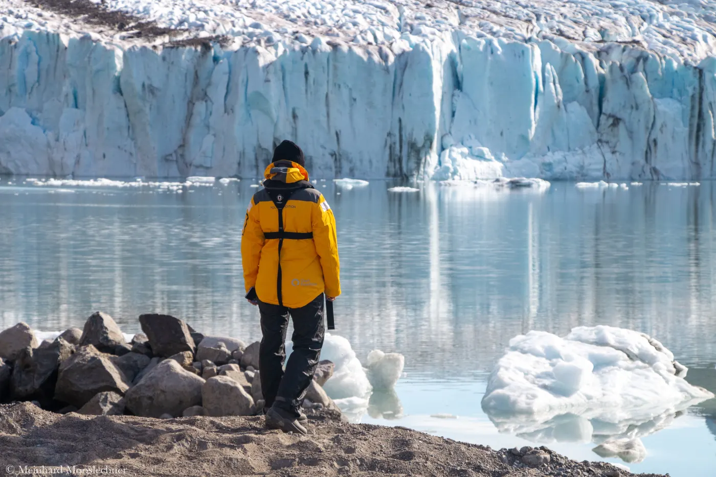 A traveller in an orange expedition jacket stands on the shore in front of a glacier front in Greenland